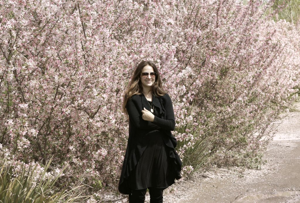 Business Mentor and Design Expert Clarissa Grace of Waking Up in Paris. She is wearing all black, photographed in front of a large bush of faint pink blossoms. 