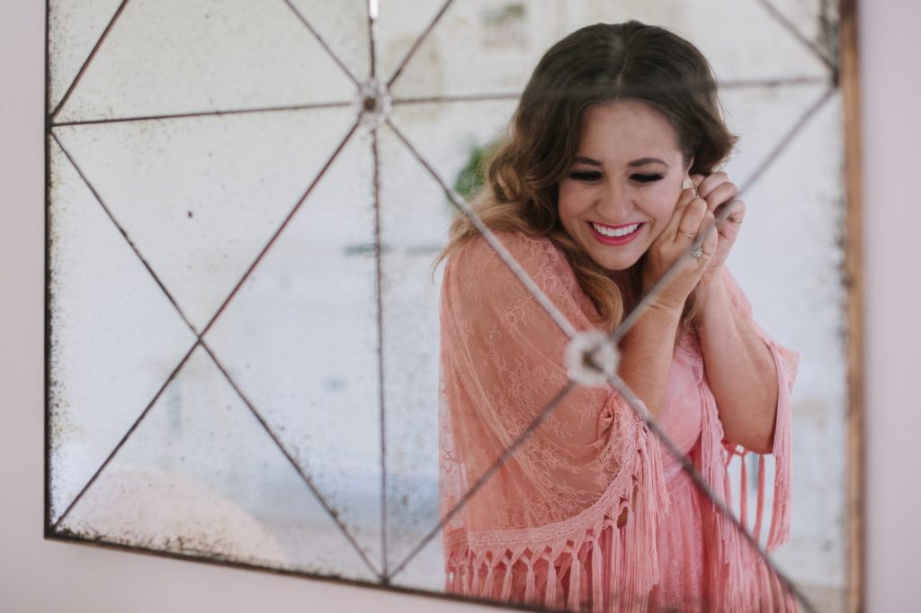 A fun photo of business mentor and design expert Clarissa Grace of Waking Up in Paris. We see Clarissa's smiling reflection in a mirror, as she puts on an earring. She is glowing in a a pink shawl and dress. 